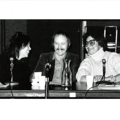 Photographic Materials depicts: black and white photo of (from left to right) May Stevens, Conrad Atkinson, Ingrid Sischy sitting at table